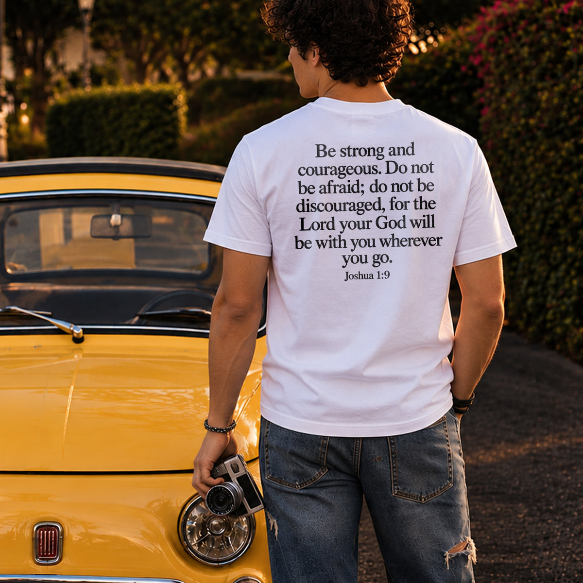 White Joshua 1:9 Christian T-shirt from Mirror Scripture Tees featuring a Bible verse print on the back, modeled beside a yellow vintage car in a lifestyle photo