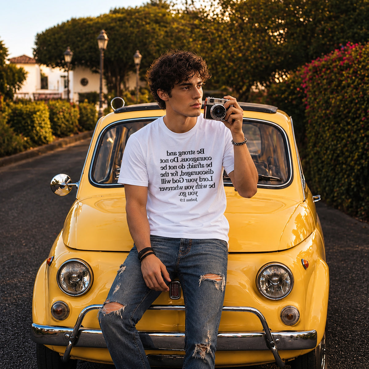 White Joshua 1:9 Christian T-shirt from Mirror Scripture Tees featuring mirrored Bible verse text on the front, modeled by a man sitting on a yellow vintage car in a lifestyle photo.